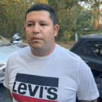 A man with short dark hair wearing a white Levi's t shirt stands outdoors in front of parked cars and trees with autumn foliage
