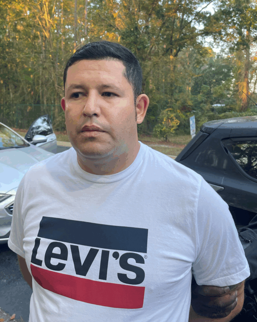 A man with short dark hair wearing a white Levi's t shirt stands outdoors in front of parked cars and trees with autumn foliage