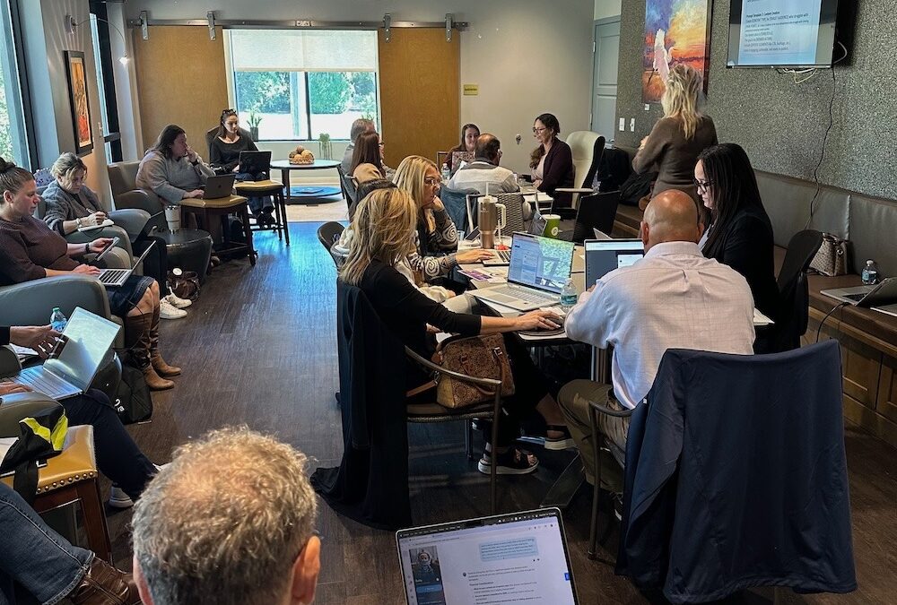 People sitting at tables in a conference room, working on laptops and taking notes A presenter stands at the front near a screen displaying a presentation The room is well lit with large windows and modern hanging lights