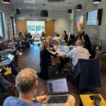 People sitting at tables in a conference room, working on laptops and taking notes A presenter stands at the front near a screen displaying a presentation The room is well lit with large windows and modern hanging lights