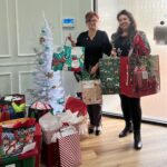 Two women stand indoors next to a small white Christmas tree, smiling and holding large gift bags More colorful gift bags and presents are stacked on the floor around them against a pale green wall