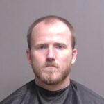 A man with short light brown hair, a beard, and a serious expression wears a dark cape against a plain gray background in a mugshot style photograph