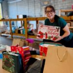 A woman with glasses smiles while holding a festive "Happy Holidays" gift bag in a room filled with various holiday bags and presents