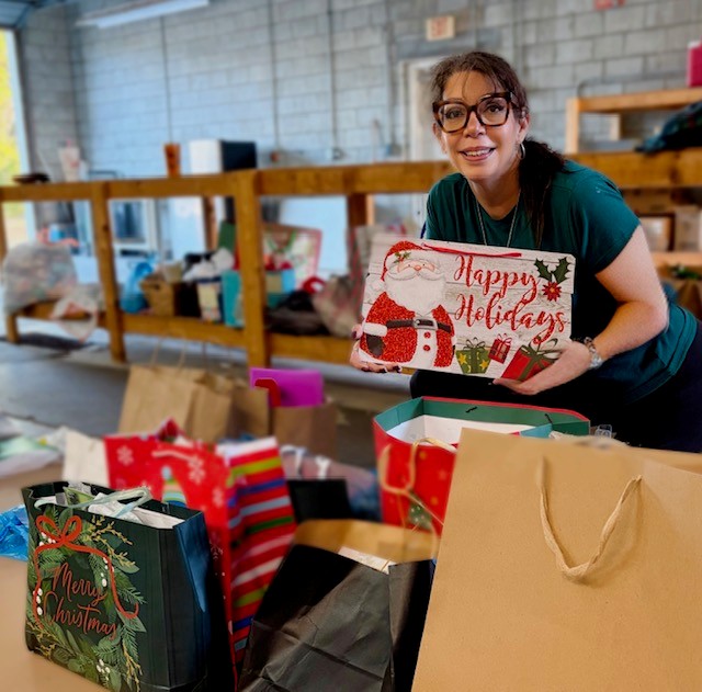 A woman with glasses smiles while holding a festive "Happy Holidays" gift bag in a room filled with various holiday bags and presents