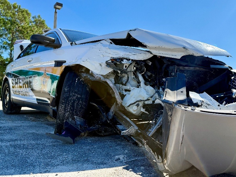 A sheriff’s patrol car with severe front end damage is parked on a paved surface, showing crumpled metal and exposed internal components Trees and a streetlight are visible in the background
