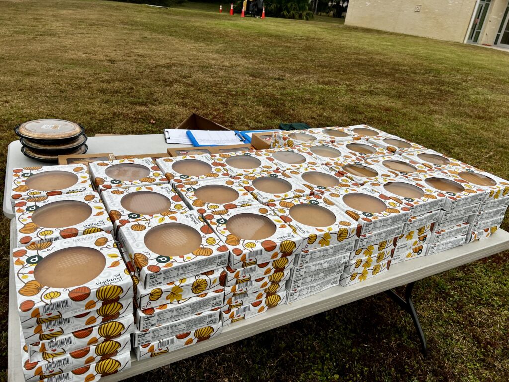 A long table outdoors holds stacks of boxed pies in festive autumn themed packaging, with several whole pies in tins and some papers or clipboards nearby The table is set on grass near a building
