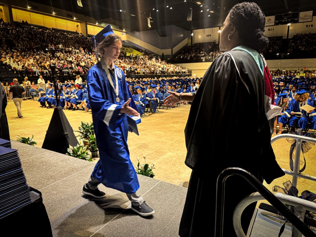 A graduate in a blue cap and gown walks across a stage to shake hands with a faculty member during a graduation ceremony, with an audience and other graduates seated in the background
