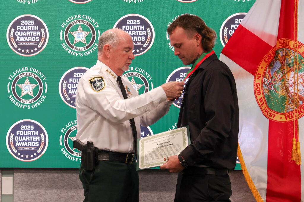 A sheriff places a medal around a man's neck during an awards ceremony The man holds a certificate, and a Florida state flag is visible in the background, along with several awards banners