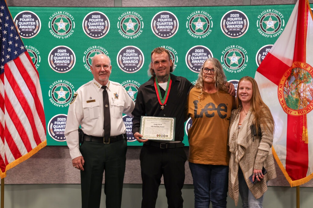 Four people stand in front of a backdrop with "Fourth Quarter Awards" and sheriff logos One man in uniform, another holds a certificate and medal, flanked by two women, all smiling American and Florida flags are visible