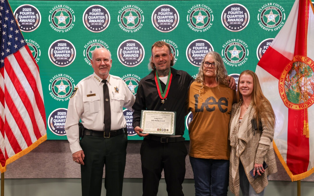 Four people stand in front of a backdrop with "Fourth Quarter Awards" and sheriff logos One man in uniform, another holds a certificate and medal, flanked by two women, all smiling American and Florida flags are visible