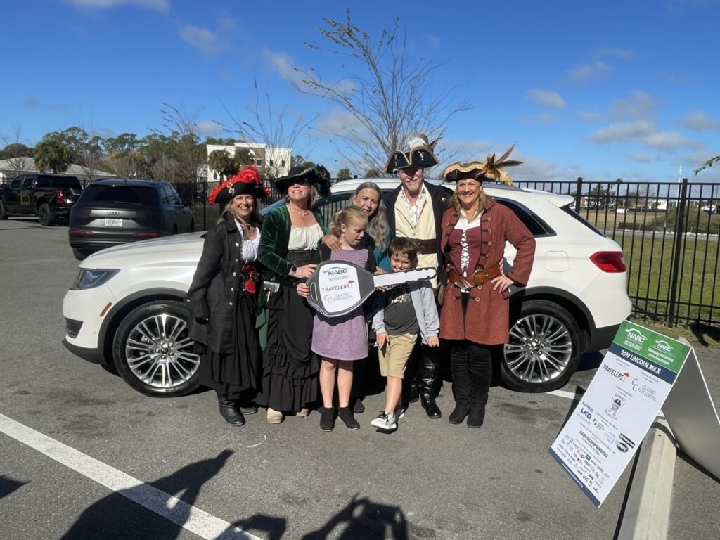 A group of people in pirate costumes stand smiling in front of a white car in a parking lot, with two children holding an oversized key A sign with sponsors’ logos is displayed nearby A group of people in pirate costumes stand smiling in front of a white car in a parking lot, with two children holding an oversized key A sign with sponsors’ logos is displayed nearby