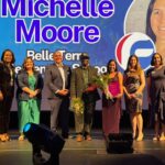 A group of ten people stand on stage, some holding flowers and plaques Behind them is a screen displaying “Michelle Moore” with her photo and text for Belle Terre Elementary School The group smiles for the picture