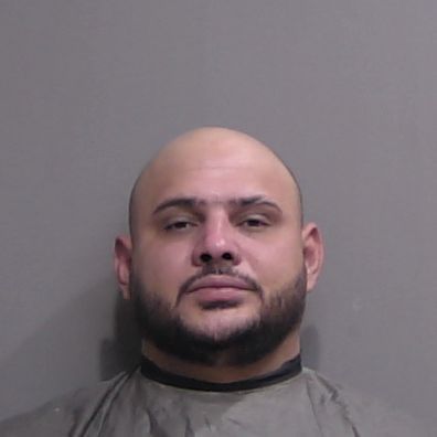 A man with a shaved head, trimmed beard, and mustache is shown in a neutral expression against a plain background He is wearing a gray garment, and the image resembles a mugshot A man with a shaved head, trimmed beard, and mustache is shown in a neutral expression against a plain background He is wearing a gray garment, and the image resembles a mugshot