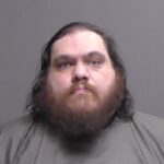 A man with long dark hair and a thick beard looks straight at the camera with a neutral expression, wearing a plain dark shirt, against a plain gray background