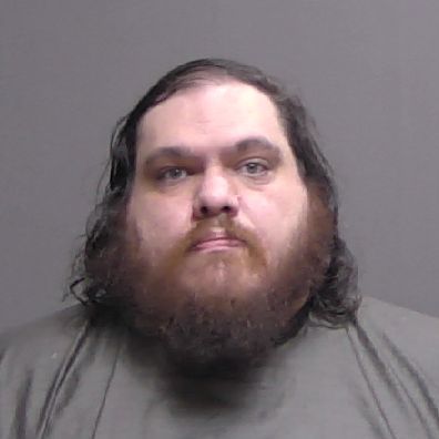 A man with long dark hair and a thick beard looks straight at the camera with a neutral expression, wearing a plain dark shirt, against a plain gray background