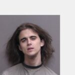 A young person with long brown hair and a faint goatee looks at the camera with a neutral expression, standing against a plain gray background