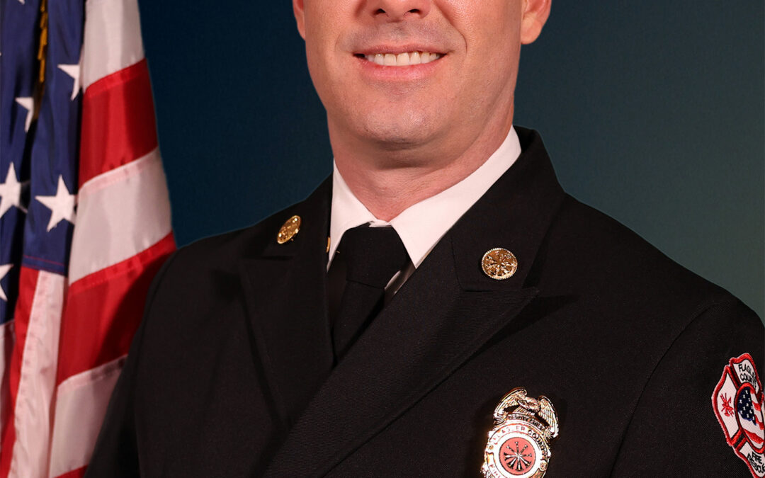 A man in a formal dark firefighter uniform with pins and a badge stands smiling in front of a U S flag, posing for a professional portrait against a dark blue background