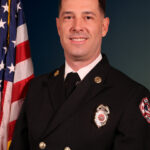 A man in a formal dark firefighter uniform with pins and a badge stands smiling in front of a U S flag, posing for a professional portrait against a dark blue background