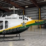 A Flagler County Sheriff's helicopter with green, yellow, and white markings is parked inside a spacious, empty hangar with exposed steel beams and large doors