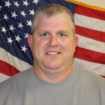A man with short light hair and a gray shirt smiles in front of a large American flag