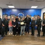 A group of people, including police officers, school staff, and students, stand together smiling indoors One person in the center holds a certificate Framed photos and a blue school emblem are visible on the wall behind them