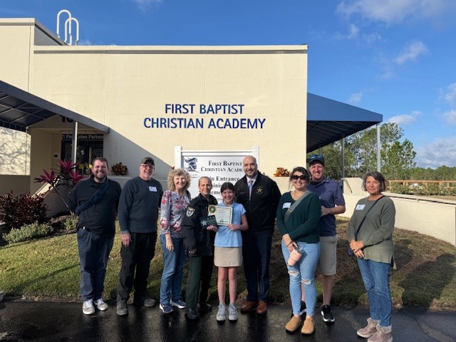 A group of nine people stand smiling in front of a building labeled “FIRST BAPTIST CHRISTIAN ACADEMY ” One person in the center holds a certificate, and everyone is posing for the photo outside on a sunny day