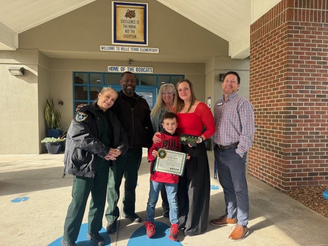 Six adults and one child stand smiling outside a school entrance The child in front holds a certificate A uniformed officer is at left A sign above the door reads “Home of the Bobcats ”