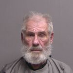 An older man with a white beard and gray hair is wearing a gray shirt and sitting against a plain gray background