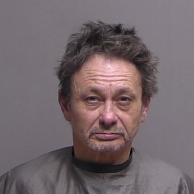 A middle aged man with tousled dark hair and a short beard wearing a gray smock poses for a mugshot against a plain, light colored background