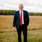A man in a dark blue suit with a red striped tie stands smiling in an open grassy field with trees and a partly cloudy sky in the background