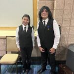 Two young people stand indoors wearing white shirts, black vests, and black pants One has a medal pinned to their vest They are smiling, with a chair and a trash bin nearby against a stone textured wall