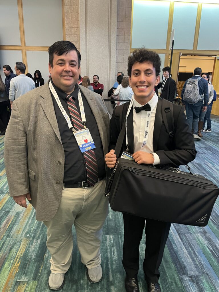 Two men are smiling at an indoor event The man on the left wears a gray suit and striped tie, while the man on the right wears a black tuxedo and holds a large black bag People are gathered and talking in the background
