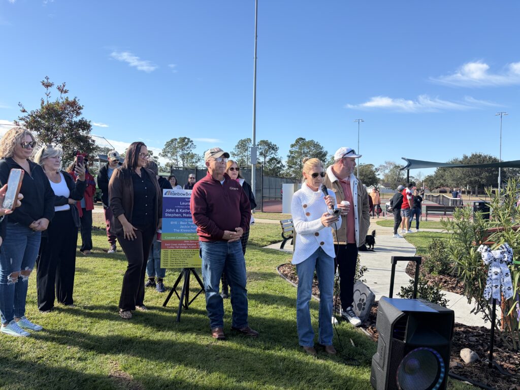 A group of people stand outdoors on a grassy area near a park A woman in a white sweater speaks into a microphone while others listen A colorful sign and speaker are nearby, with trees and a sports field in the background