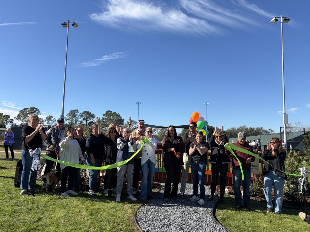 A group of people stands outdoors on grass under a blue sky, smiling and holding a green ribbon for a ribbon cutting ceremony Some hold balloons, and several are making hand signs There are trees and lights in the background