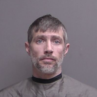 A man with short brown and gray hair, a short beard, and blue eyes poses against a plain gray background, wearing a dark cape like garment similar to those worn in mugshots A man with short brown and gray hair, a short beard, and blue eyes poses against a plain gray background, wearing a dark cape like garment similar to those worn in mugshots