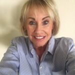 A smiling older woman with short blonde hair wears a blue and white striped button up shirt and stands in front of a plain light colored wall