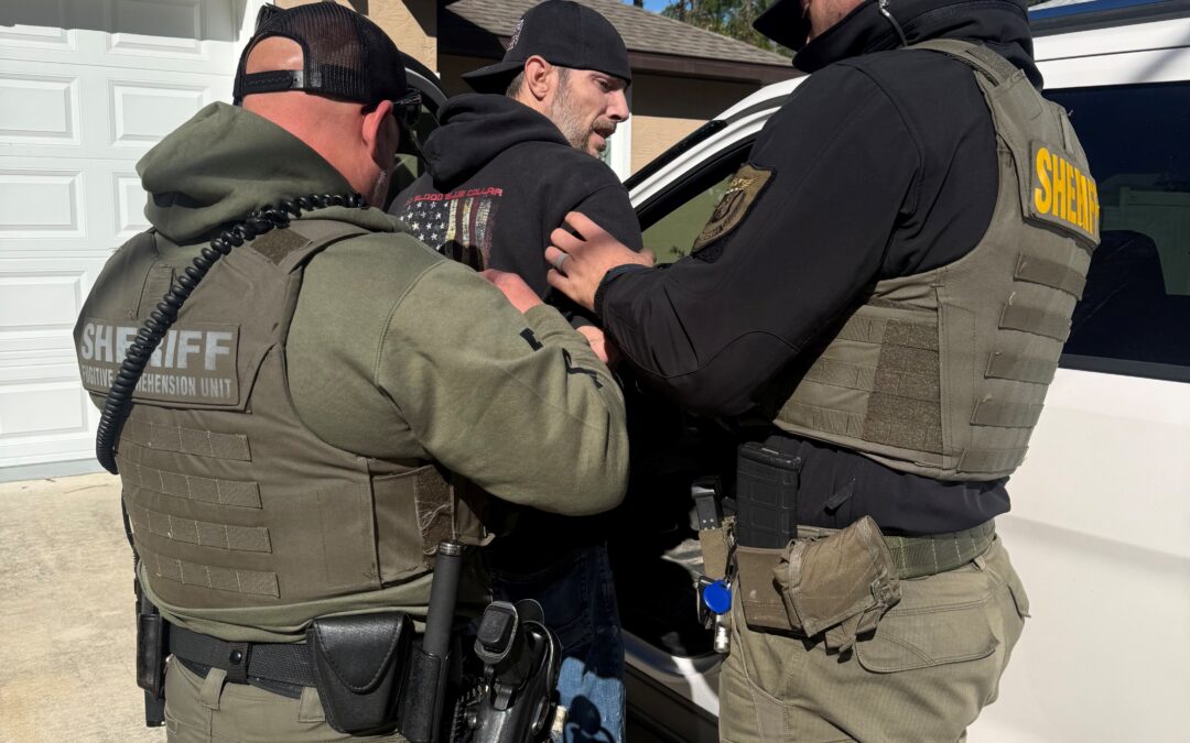 Two sheriff officers in tactical gear detain and handcuff a man in a black hoodie next to a white vehicle in a suburban driveway, with pine trees and a house in the background