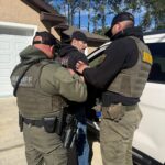 Two sheriff officers in tactical gear detain and handcuff a man in a black hoodie next to a white vehicle in a suburban driveway, with pine trees and a house in the background