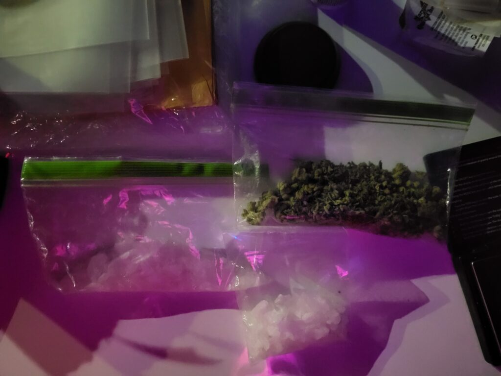 Two clear plastic bags on a surface, one containing green leafy material and the other containing white crystalline chunks, both illuminated by purple tinted light Other containers and papers are nearby