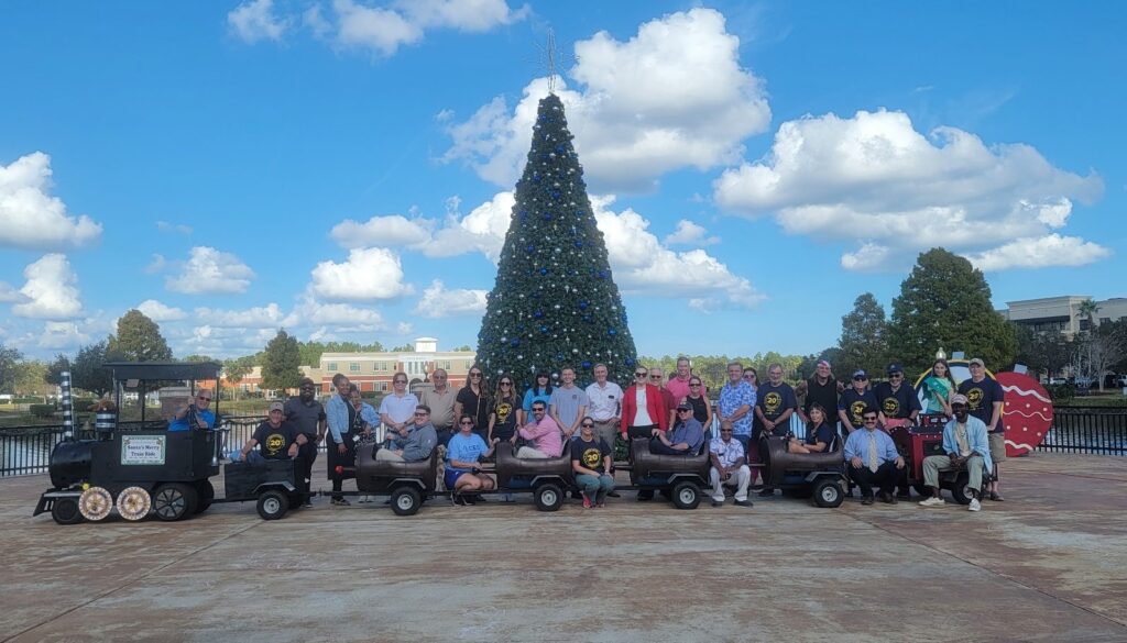 A large group of people pose in front of a decorated Christmas tree outdoors, with blue sky and clouds overhead They are sitting and standing around a small black train with carts on a pavement near a park or waterfront