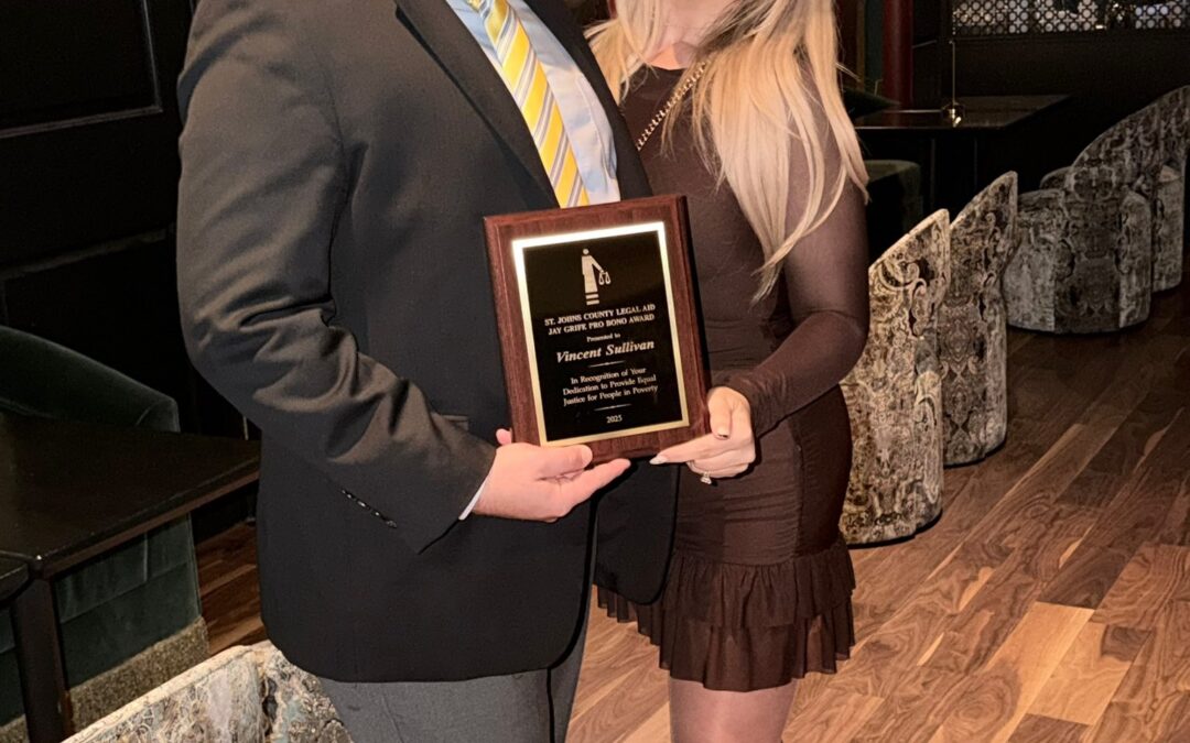 A man in a suit holding a plaque stands next to a woman in a brown dress Both are smiling, with the woman’s arm around the man They are in an elegant venue with art and patterned seating in the background