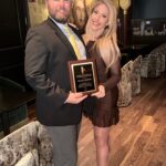 A man in a suit holding a plaque stands next to a woman in a brown dress Both are smiling, with the woman’s arm around the man They are in an elegant venue with art and patterned seating in the background