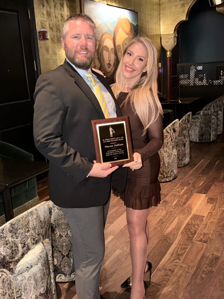 A man in a suit holding a plaque stands next to a woman in a brown dress Both are smiling, with the woman’s arm around the man They are in an elegant venue with art and patterned seating in the background