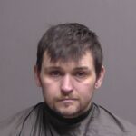 A man with short dark hair and facial stubble is wearing a dark colored drape, posing against a plain gray background