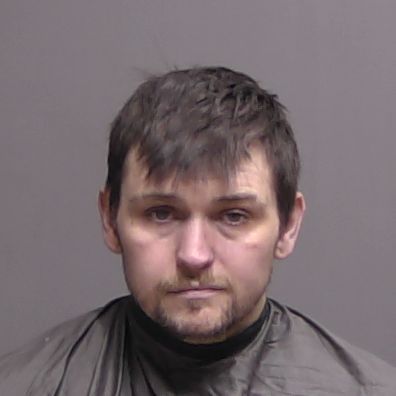 A man with short dark hair and facial stubble is wearing a dark colored drape, posing against a plain gray background