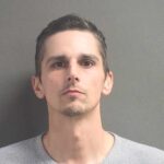 A man with short dark hair, light skin, and a trimmed goatee is wearing a light gray henley shirt and standing against a plain gray background, looking directly at the camera with a neutral expression