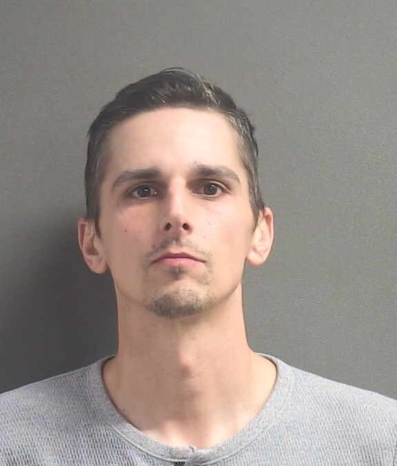 A man with short dark hair, light skin, and a trimmed goatee is wearing a light gray henley shirt and standing against a plain gray background, looking directly at the camera with a neutral expression