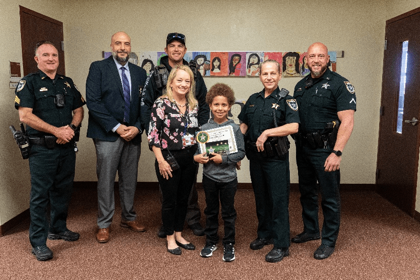 A child holds a certificate and stands with two women and four uniformed officers, all smiling in a hallway with student artwork displayed on the wall behind them