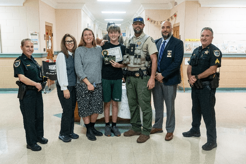 A group of seven people, including three uniformed officers, stand smiling in a school hallway One teenager in the center holds a certificate, flanked by adults in casual and professional attire
