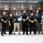 A group of eight people, including law enforcement officers and civilians, stand in front of a trophy case One young man in the center holds a certificate and poses with others smiling beside him